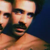 Double-exposure glitch portrait of a shirtless man with curly dark hair and a mustache-goatee, gazing off to the side against a deep blue background; his image is duplicated and offset to the left with a cyan/red chromatic distortion effect, and a thin gold chain is visible around his neck.